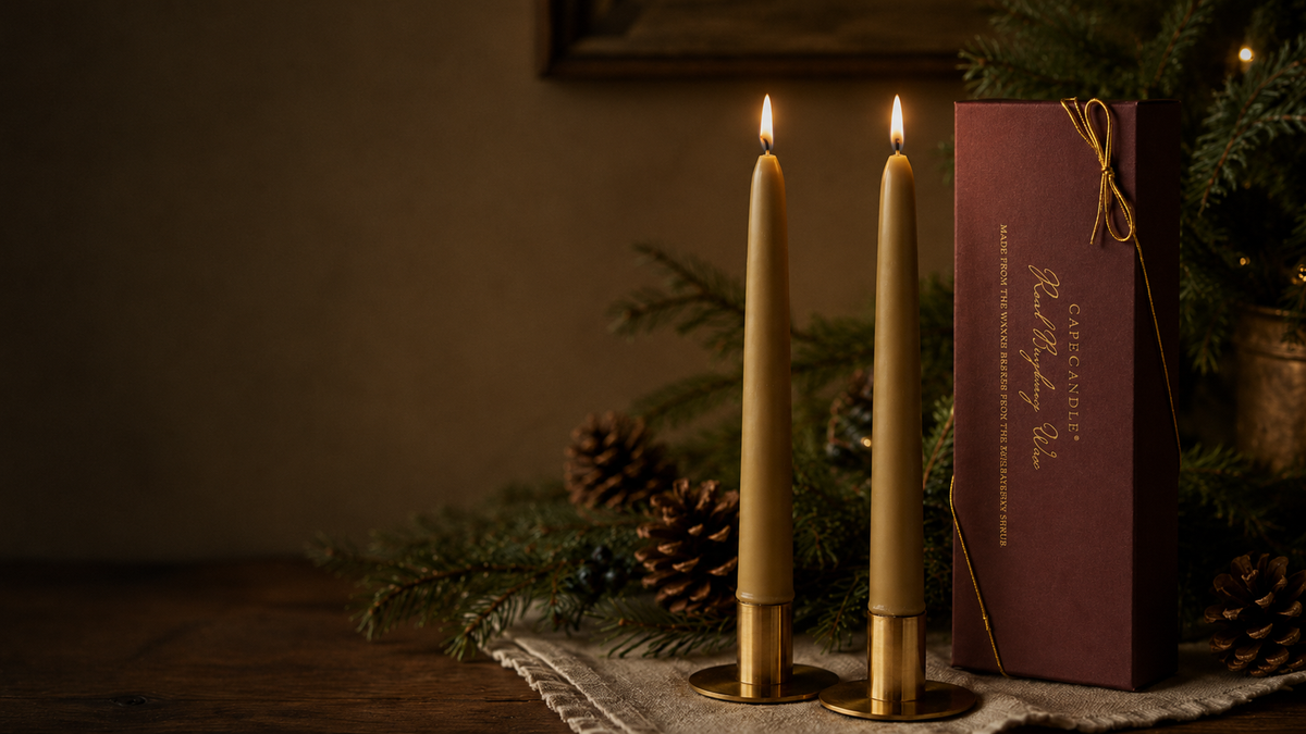 Two lit candles in gold holders with a box labeled 'Candle' on a textured surface with greenery.